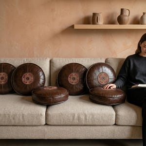 Può includere: Una collezione di cuscini rotondi in pelle marrone con motivi centrali decorativi in rosso, arancione e crema. I cuscini sono disposti su un divano beige, creando un'area salotto calda e invitante. I cuscini variano in dimensioni.