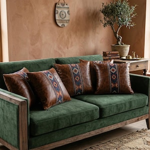 Könnte beinhalten: Ein grünes Sofa mit vier braunen Lederkissen. Jedes Kissen hat ein zentrales gemustertes Band mit blauen, orangefarbenen und weißen geometrischen Designs. Das Sofa hat einen Holzrahmen und befindet sich in einem Raum mit einem dekorativen Wandbehang.