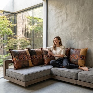 May include: A modern sectional sofa with gray cushions and a wooden frame. The sofa is adorned with four decorative leather pillows. The pillows feature a patchwork design with brown, tan, and black leather, accented with colorful embroidered patterns.