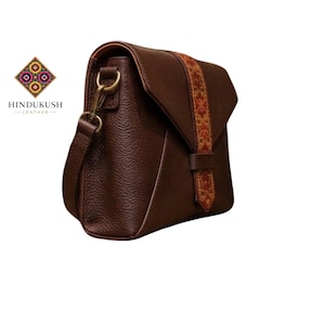 Könnte beinhalten: Braune Leder-Umhängetasche mit strukturierter Oberfläche. Die Tasche hat einen dekorativen Riemen mit farbenfrohem geometrischen Muster. Sie hat einen Überschlagverschluss und einen langen, verstellbaren Riemen. Das Markenlogo "HINDUKUSH LEATHER" ist sichtbar.