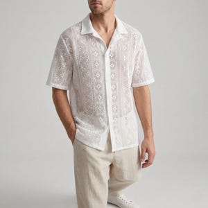Puede incluir: Camisa blanca de manga corta con cuello abierto y estampado geométrico. La camisa está abotonada por delante y tiene un corte holgado. El modelo lleva pantalones beiges y zapatillas blancas.