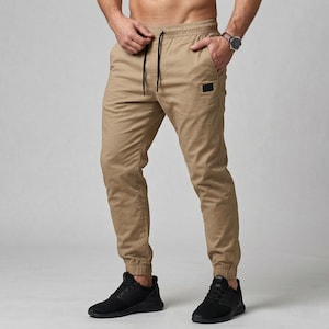 May include: Khaki-colored jogger pants with a drawstring waist and elastic cuffs. The pants have a small black square patch near the pocket and are paired with black athletic shoes. The model is wearing a watch.