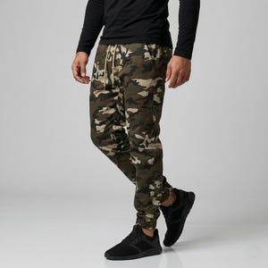 May include: Camouflage print joggers with a drawstring waist and elastic cuffs. The pants feature a pattern of green, brown, and tan. The person is wearing black sneakers and a black long-sleeve shirt.