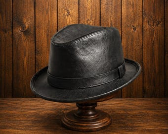 Pode incluir: Um chapéu fedora preto com um acabamento liso e texturizado, apresentando uma faixa preta a condizer em torno da base da copa. O chapéu possui um topo vincado e uma aba estruturada, exibindo um design clássico com costuras visíveis.