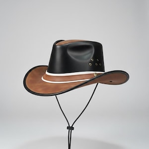 Puede incluir: Un sombrero de vaquero de cuero con copa negra y ala marrón. El sombrero presenta una franja blanca y una correa para la barbilla. El sombrero tiene un emblema dorado en el lateral y está diseñado para la protección solar.