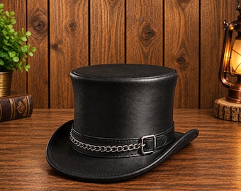 May include: A black top hat featuring a flat crown and a curved brim. The hat is adorned with a black band that includes a silver-toned metal buckle and a decorative silver-toned chain link detail wrapped around the base of the crown.