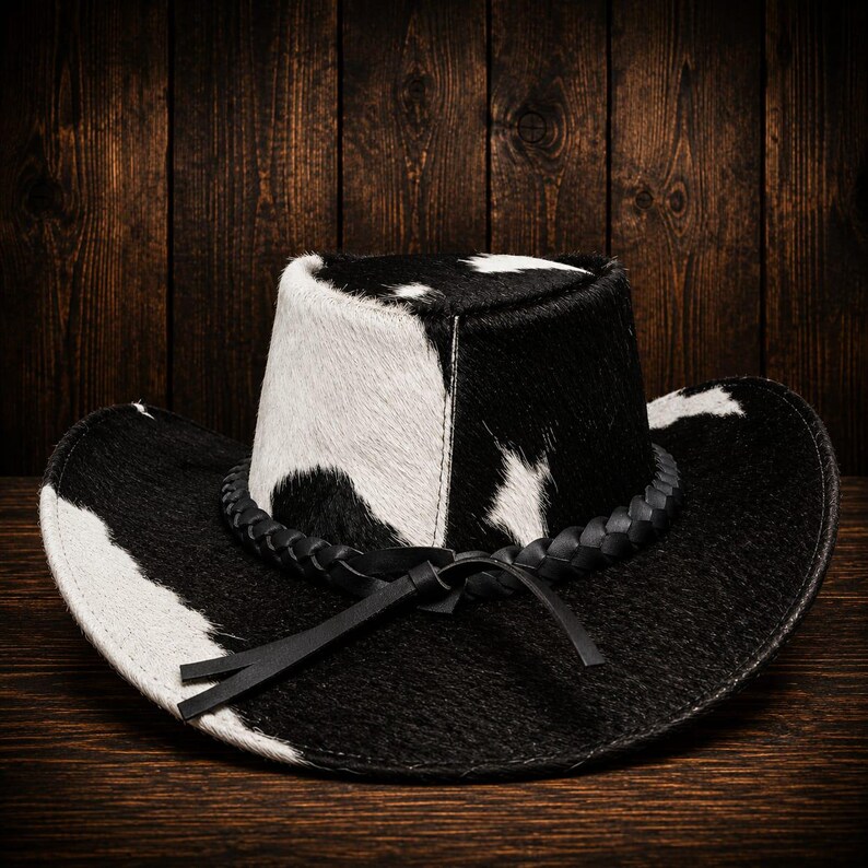 May include: A cowboy hat featuring a black and white cow print pattern with a textured hair-on-hide finish. The hat includes a black braided leather band around the base of the crown, which is tied in a simple knot at the front of the brim.