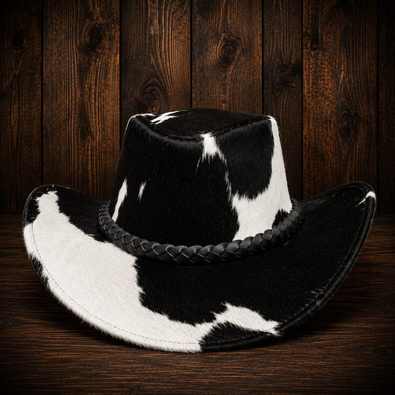 May include: A cowboy hat featuring a black and white cow print pattern with a textured, hair-on-hide appearance. The hat includes a black braided band encircling the base of the crown, which has a flat top and a wide, curved brim.
