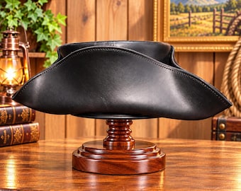 May include: A black tricorn hat with a smooth finish, featuring a wide, curved brim that is folded up on three sides. The hat is displayed resting on a polished, round, tiered wooden stand with a dark brown grain finish.