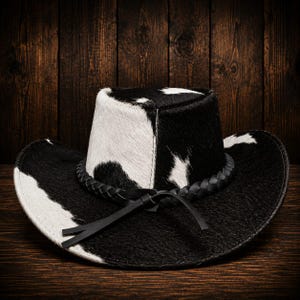 May include: A cowboy hat featuring a black and white cow print pattern with a textured hair-on-hide finish. The hat includes a black braided leather band around the base of the crown, which is tied in a simple knot at the front of the brim.