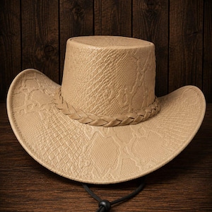 May include: Tan cowboy hat with a snakeskin pattern and a braided band around the crown. The hat has a wide brim and a black chin strap. The hat is made of a textured material and sits on a wooden surface.