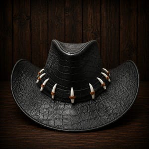 May include: A black cowboy hat featuring a textured crocodile skin pattern across the crown and wide brim. A black band encircles the base of the crown, decorated with a series of white, curved, tooth-like accents secured by brown leather ties.