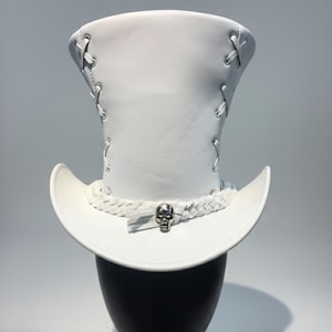 May include: A white top hat featuring a tall, structured crown with side lacing details. The base of the crown is encircled by a braided white band, which is accented at the front by a polished silver-toned skull emblem with a small tassel.
