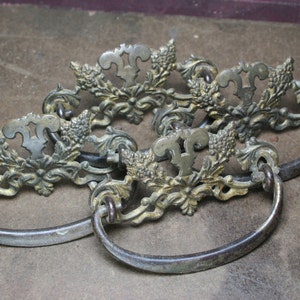 May include: Three antique brass drawer pulls with ornate floral and leaf designs. Each pull has a loop handle.