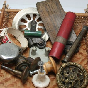 May include: A wicker basket filled with various vintage hardware items, including a metal measuring tape, a red and gold cylinder, a brass door knob, a metal wheel, and a green and silver plumbing part.