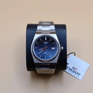 Op de afbeelding: Een zilveren Tissot PRX Powermatic 80 horloge met een blauwe wijzerplaat en een roestvrijstalen band. Het horloge wordt getoond op een zwarte standaard met een wit label eraan. De wijzerplaat heeft het Tissot-logo en de tekst "1853".