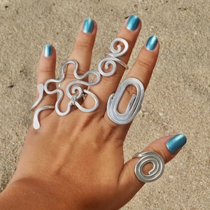 May include: Four silver-toned wire rings worn on a hand. One ring features a complex, multi-loop floral-like design, another is a simple spiral, one is a vertical oval shape, and the last is a small spiral on the thumb. All have a metallic finish.