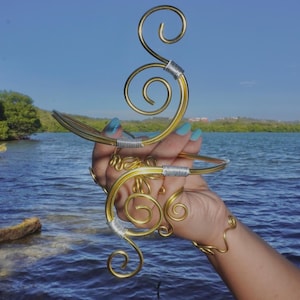 May include: A gold-colored wire arm cuff featuring intricate spiral designs and silver-toned wire wrappings at the connection points. The metal structure is formed into multiple circular coils and curved lines, creating a decorative piece of jewelry.