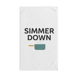 Op de afbeelding: Witte keukendoek met de tekst "SIMMER DOWN" in zwarte letters. Daaronder een teal-kleurige pan met stoom en een bruin handvat. De handdoek is rechthoekig.