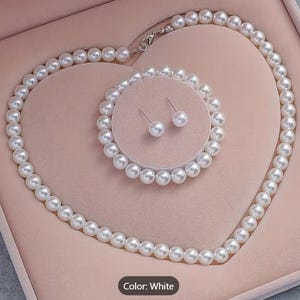Op de afbeelding: Een sieradenset bestaande uit een witte parelketting, een bijpassende witte parelarmband en twee witte parelknopjes als oorbellen. De ketting is voorzien van een zilverkleurige metalen sluiting. De items liggen in een hartvormig doosje.