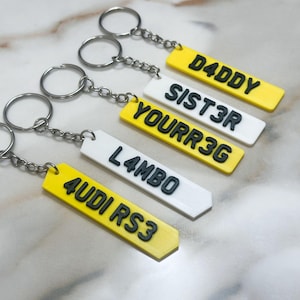 May include: Five rectangular keychains with metal rings and chains. Three are yellow with black raised text reading D4DDY, YOURR3G, and 4UDI RS3. Two are white with black raised text reading SIST3R and L4MBO. Each keychain has a pointed end.