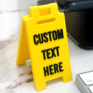 May include: A bright yellow A-frame sign with a hinged top, featuring the black raised text CUSTOM TEXT HERE on the front panel. The sign is constructed with a smooth, matte finish and stands upright on its own four feet in a triangular shape.
