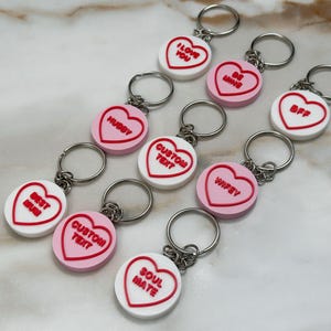 May include: A collection of round keychains featuring heart-shaped designs in white and pink. Each keychain has a metal ring and red embossed text including I LOVE YOU, BE MINE, BFF, HUBBY, CUSTOM TEXT, WIFEY, BEST MUM, and SOUL MATE.