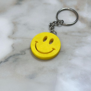 Puede incluir: Un llavero circular de color amarillo brillante con un diseño clásico de cara sonriente con dos ojos ovalados y una boca curva. La cara sonriente amarilla está unida a una anilla de metal plateado mediante una cadena corta de pequeños eslabones.
