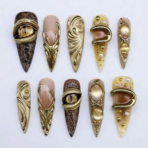 May include: A set of ten stiletto-shaped press-on nails featuring gold metallic accents, sculpted relief patterns, and realistic dragon eye designs. The nails incorporate a colour palette of nude, brown, and gold with small white pearl embellishments.