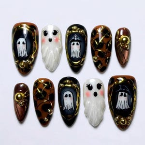 May include: A set of ten press-on nails featuring a mix of designs including white ghosts with pink cheeks, brown and black tortoiseshell patterns, and gold metallic accents. The nails are almond-shaped with glossy finishes and raised gold beads.