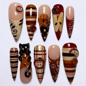 May include: A set of ten press-on nails featuring autumn-themed designs in shades of brown, cream, and red. The nails include patterns of stripes, plaid, swirls, and 3D embellishments such as small buttons, a pumpkin, an apple, and a black cat.