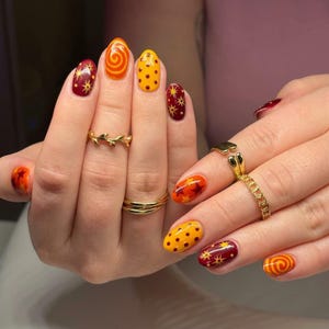 May include: Oval-shaped fingernails featuring a mix of designs including orange swirls, yellow with red polka dots, and deep red with gold starburst patterns. Three gold-toned metal rings are worn on the fingers, including a leaf-shaped band.