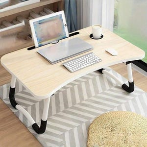 May include: A light wood-effect laptop desk with a curved edge, featuring a built-in tablet holder, cup holder, and a slot for a laptop. The desk has white legs with black accents and is set on a grey and white patterned rug. A laptop, keyboard, and mouse are on the desk.