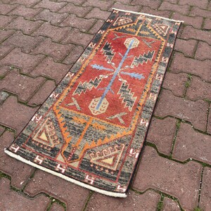 Puede incluir: Una alfombra rectangular tejida a mano con un campo central rojo y patrones geométricos intrincados en negro, naranja y beige. Un diseño azul en forma de cruz está centrado en la alfombra. La alfombra tiene flecos blancos en ambos extremos.