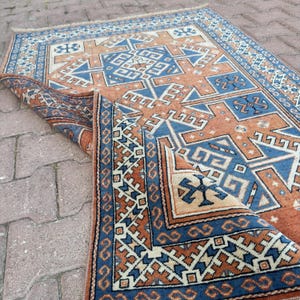 Könnte beinhalten: Ein rechteckiger Teppich mit geometrischem Muster in Rostorange, Creme und Dunkelblau. Das Design umfasst komplexe Bordüren und zentrale Motive, wobei eine umgeschlagene Ecke die Unterseite des gewebten Textilstoffs freilegt.