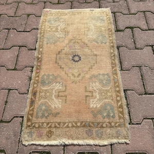 Puede incluir: Una alfombra tejida rectangular con un fondo color melocotón descolorido y un medallón central de diamante en azul oscuro, rodeado de patrones geométricos en verde apagado y bronceado, con flecos blancos cortos en los bordes.