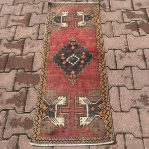 Puede incluir: Una alfombra rectangular de estilo vintage con un campo central rojo. La alfombra presenta patrones geométricos en marrón, beige y naranja. El diseño incluye una forma de diamante central y motivos en forma de cruz en cada extremo. La alfombra probablemente está hecha de material tejido.