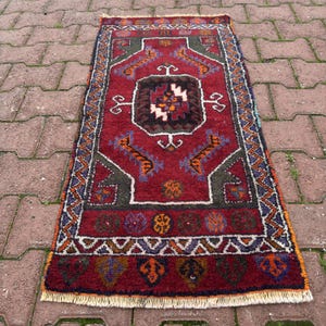 Puede incluir: Una alfombra rectangular, tejida a mano, con un fondo rojo oscuro y patrones geométricos intrincados. La alfombra presenta un medallón central con un diseño complejo en tonos marrones, blancos y naranjas, rodeado de bordes verdes, azules y naranjas. La alfombra tiene un borde con flecos.