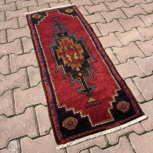 May include: A rectangular red rug with a central geometric design in black, gold, and blue. The rug has a black border with decorative elements and a white fringe on the ends. The rug is approximately 3 feet long.