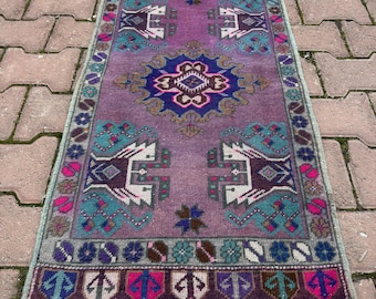 Könnte beinhalten: Ein rechteckiger Teppich mit violettem Grund und einem zentralen geometrischen Medaillon in Blau und Pink. Der Teppich zeigt symmetrische, stilisierte Tierfiguren in Weiß und Dunkelviolett, umrahmt von floralen Mustern und Fransen.