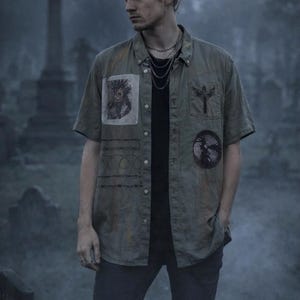 May include: Olive green short-sleeve button-down shirt with multiple patches and a black undershirt. The patches include a tribal design, a cross with wings, and a circular graphic. The person is wearing a silver chain necklace and dark pants.