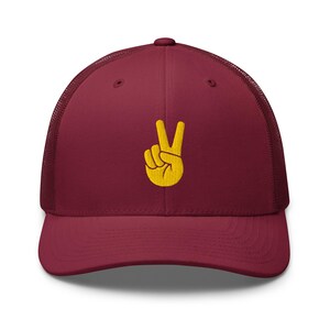 May include: A maroon trucker hat featuring a mesh back and a solid front panel. Centered on the front is an embroidered yellow graphic of a hand making a peace sign gesture with the index and middle fingers extended upward.