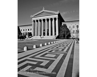 May include: A black and white photograph of a large building with columns and a patterned walkway in front. The building has a large entrance with a set of double doors. The walkway is made of black and white tiles and leads to the building.