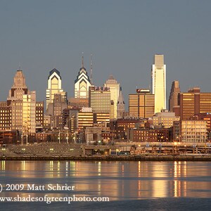 May include: A panoramic view of the Philadelphia skyline at dusk, with the city's iconic skyscrapers illuminated against a twilight sky. The buildings are reflected in the calm waters of the Delaware River.
