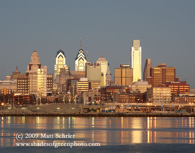May include: A panoramic view of the Philadelphia skyline at dusk, with the city's iconic skyscrapers illuminated against a twilight sky. The buildings are reflected in the calm waters of the Delaware River.