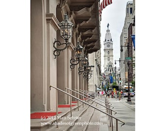 May include: A view of the Philadelphia City Hall from a building entrance with a red carpet and ornate black metal lamp posts. The building has a white facade and a series of steps leading up to the entrance.