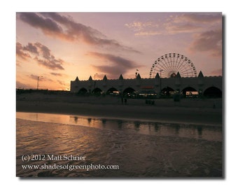 May include: A silhouette of a Ferris wheel and a building with arches against a pink and purple sunset sky. The beach is in the foreground with the water reflecting the sunset colors.