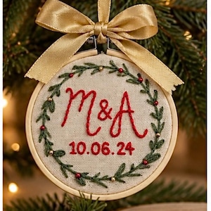 Pode incluir: Um ornamento circular em bastidor de bordado com fundo de tecido creme. O centro exibe o texto bordado em vermelho M&A acima da data 10.06.24. Uma guirlanda bordada em verde com pequenas contas vermelhas e douradas rodeia o texto.
