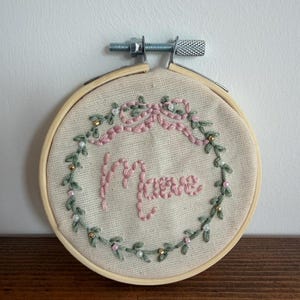 Pode incluir: Um bastidor de bordado circular de madeira contendo tecido branco sujo com bordado feito à mão. O design apresenta uma coroa de folhas verdes com pequenas contas e um laço rosa no topo. O nome Maeve está bordado em rosa no centro.
