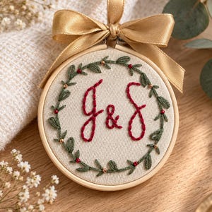 Pode incluir: Um bastidor de bordado circular de madeira com tecido cor de creme, apresentando as letras G & S bordadas em vermelho escuro no centro, rodeadas por uma coroa de folhas verdes com pequenas bagas vermelhas e contas douradas, com um laço de cetim dourado.